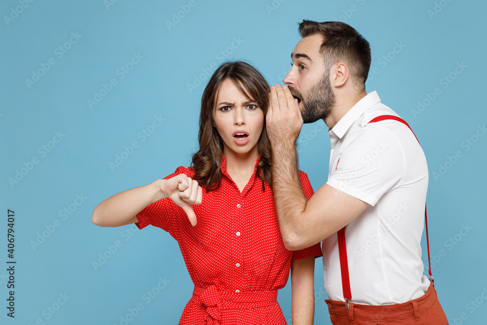 Worried young couple friends man woman in white red clothes whispering gossip tells secret behind hand showing thumb down isolated on blue background studio portrait. Valentine's Day holiday concept. Worried young couple friends man woman in white red clothes whispering gossip tells secret behind hand showing thumb down isolated on blue background studio portrait. Valentine's Day holiday concept.