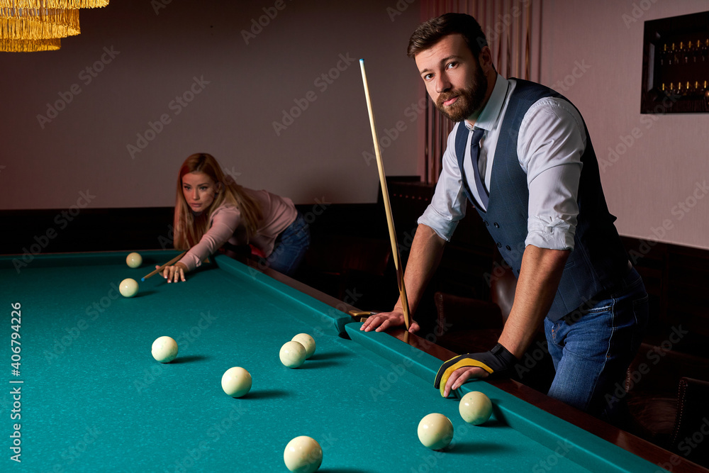 Fototapeta premium young active friends are playing billiards in bar after work, have rest and leaisure time, preparing aiming to shoot pool balls