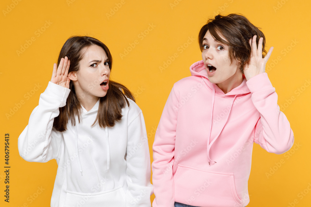 Shocked curious two young women friends 20s wearing casual white pink hoodies standing try to hear you overhear listening intently isolated on bright yellow color wall background studio portrait. Shocked curious two young women friends 20s wearing casual white pink hoodies standing try to hear you overhear listening intently isolated on bright yellow color wall background studio portrait.