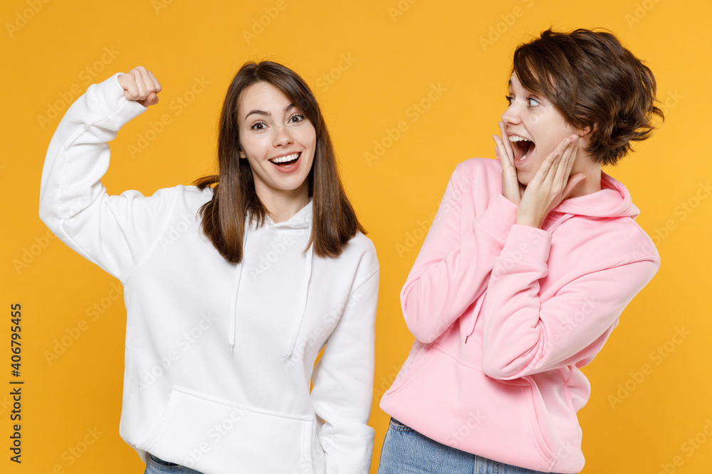 Excited sporty two young women friends 20s wearing casual white pink hoodies standing showing biceps muscles on hand put hand on cheeks isolated on bright yellow color wall background studio portrait. Excited sporty two young women friends 20s wearing casual white pink hoodies standing showing biceps muscles on hand put hand on cheeks isolated on bright yellow color wall background studio portrait.