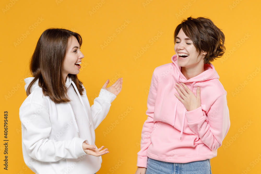Laughing funny two young women friends 20s wearing casual white pink hoodies standing speaking talking spreading hands looking at each other isolated on bright yellow color background studio portrait. Laughing funny two young women friends 20s wearing casual white pink hoodies standing speaking talking spreading hands looking at each other isolated on bright yellow color background studio portrait.