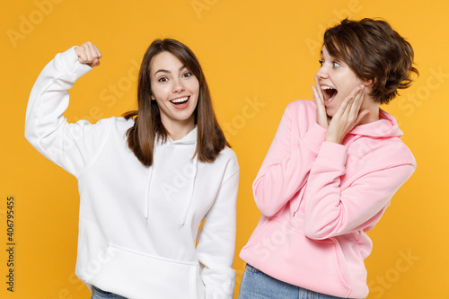 Excited sporty two young women friends 20s wearing casual white pink hoodies standing showing biceps muscles on hand put hand on cheeks isolated on bright yellow color wall background studio portrait.