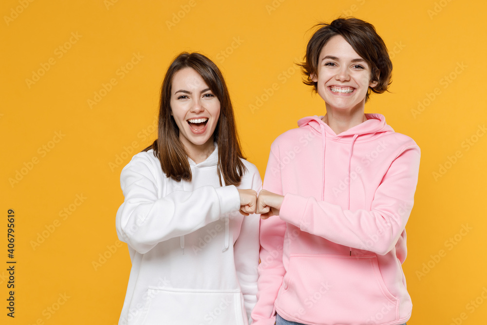 Cheerful laughing two young women friends 20s wearing casual basic white pink hoodies hold hands folded giving fists bump looking camera isolated on bright yellow color background studio portrait.
