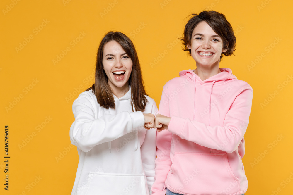 Cheerful laughing two young women friends 20s wearing casual basic white pink hoodies hold hands folded giving fists bump looking camera isolated on bright yellow color background studio portrait. Cheerful laughing two young women friends 20s wearing casual basic white pink hoodies hold hands folded giving fists bump looking camera isolated on bright yellow color background studio portrait.