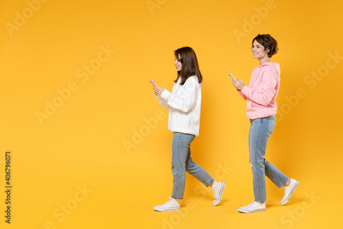 Full length side view of cheerful two young women friends 20s wearing casual white pink hoodies using mobile cell phone typing sms message isolated on bright yellow color background studio portrait.