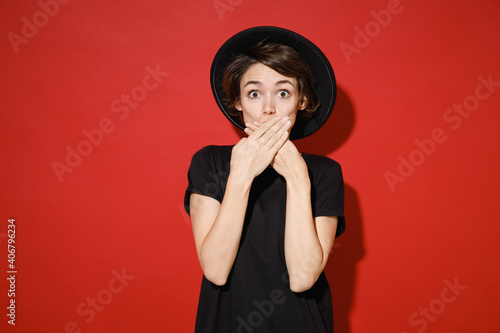 Shocked amazed worried young brunette woman 20s years old in casual basic black t-shirt hat standing covering mouth with hands looking camera isolated on bright red color background studio portrait.