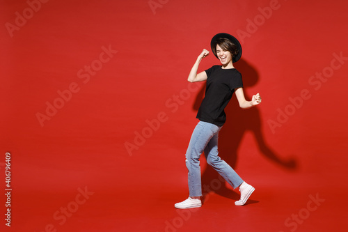 Full length side view of joyful young brunette woman 20s in casual black t-shirt hat doing winner gesture celebrating clenching fists say yes isolated on bright red color background studio portrait.