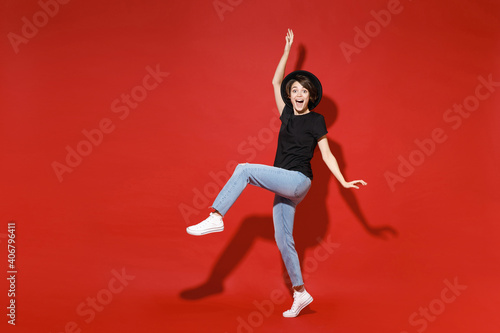 Full length side view of excited amazed cheerful young brunette woman 20s in casual basic black t-shirt hat dancing rising leg spreading hands isolated on bright red color background studio portrait.
