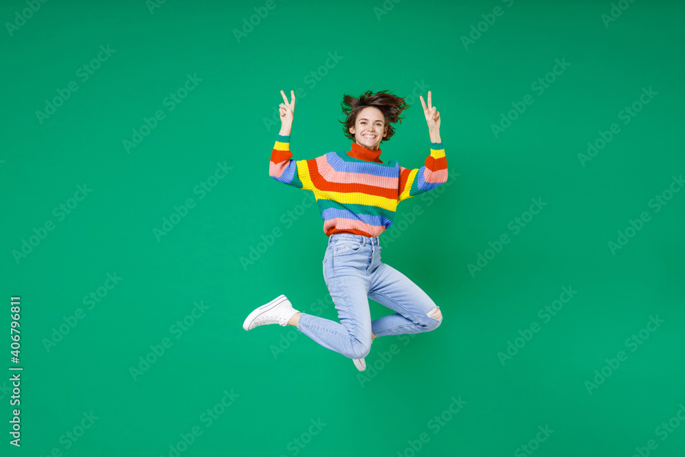 Full length of smiling cheerful funny young brunette woman 20s years old wearing basic casual colorful sweater jumping showing victory sign isolated on bright green color background studio portrait. Full length of smiling cheerful funny young brunette woman 20s years old wearing basic casual colorful sweater jumping showing victory sign isolated on bright green color background studio portrait.