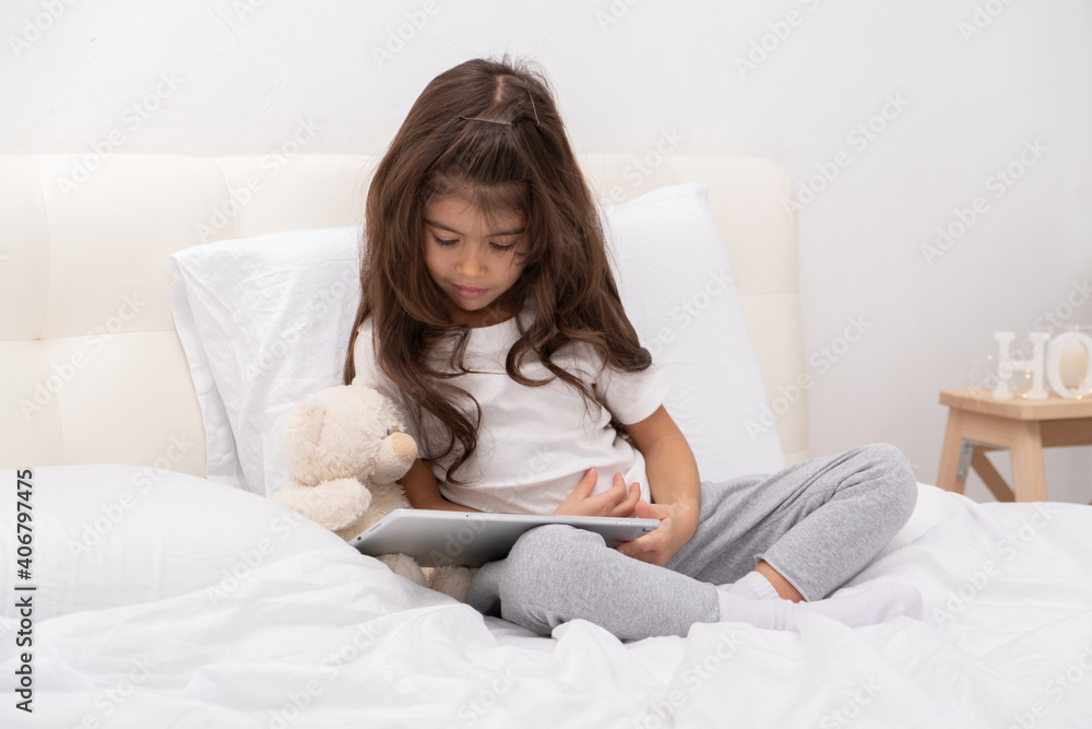 little girl in home wear with teddy bear using tablet sitting on bed little girl in home wear with teddy bear using tablet sitting on bed