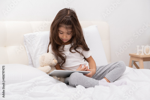 little girl in home wear with teddy bear using tablet sitting on bed