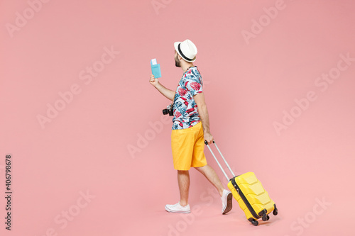 Full length side view of young traveler tourist man in summer clothes hat hold suitcase passport tickets isolated on pink color background. Passenger traveling on weekends. Air flight journey concept.