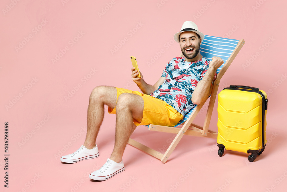 Full length happy young traveler tourist man in hat sit on deck chair using mobile cell phone doing winner gesture isolated on pink background. Passenger travel on weekend. Air flight journey concept. - obrazy, fototapety, plakaty 