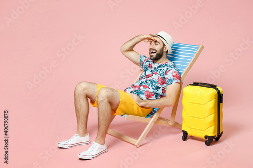 Valokuvatapetti Full length excited young tourist man in hat sit on deck chair holding hand at forehead looking far away distance isolated on pink background