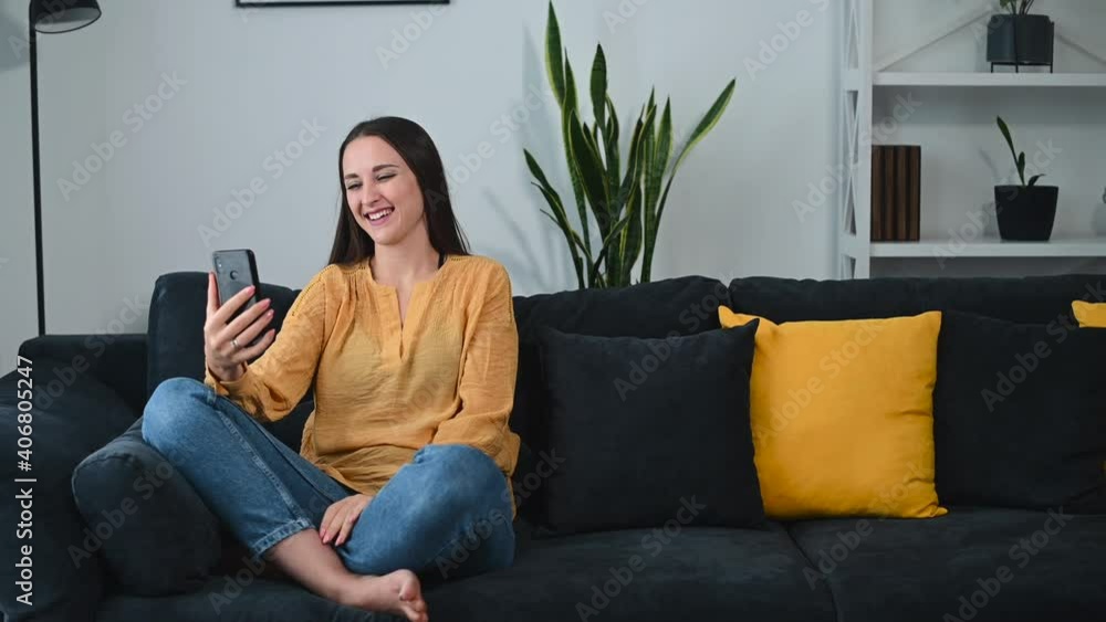 A woman meets with friends using online video call, virtual conference while staying home. She sits in relaxed pose with a smartphone on the sofa waving into webcam. Connection on the distance A woman meets with friends using online video call, virtual conference while staying home. She sits in relaxed pose with a smartphone on the sofa waving into webcam. Connection on the distance