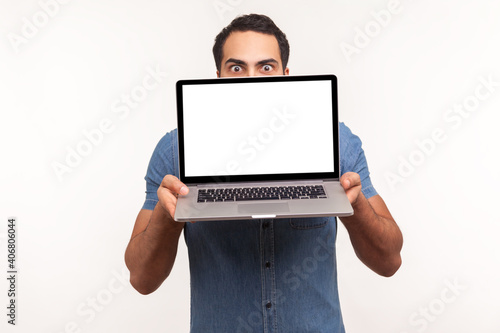 Man in blue shirt hiding behind white empty screen laptop, surprised with advertising area, looking at camera with big eyes, updating operation system. Indoor studio shot isolated on white background photo