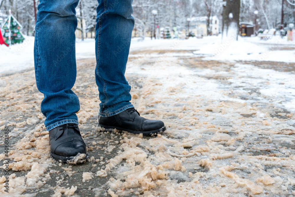 A man stands on the sidewalk in snow and mud. Black shoes and blue jeans close-up on the background of dirty snow. Ice on the road and sidewalk, the use of reagents, sand and salt.