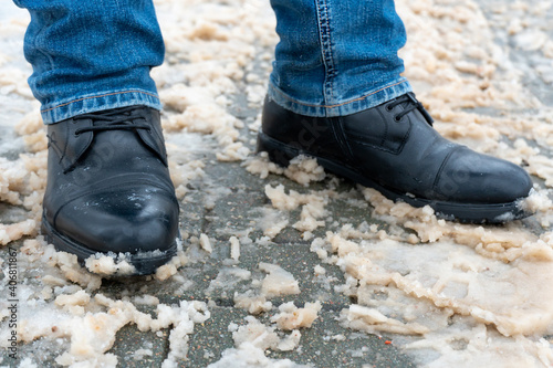 A man stands on the sidewalk in snow and mud. Black shoes and blue jeans close-up on the background of dirty snow. Ice on the road and sidewalk, the use of reagents, sand and salt.