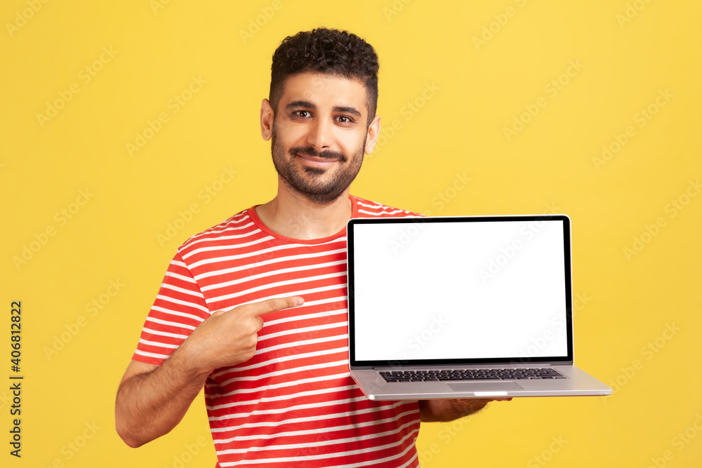 Smiling positive man with beard in striped t-shirt pointing finger at laptop with empty white display, recommending online education courses. Indoor studio shot isolated on yellow background Smiling positive man with beard in striped t-shirt pointing finger at laptop with empty white display, recommending online education courses. Indoor studio shot isolated on yellow background