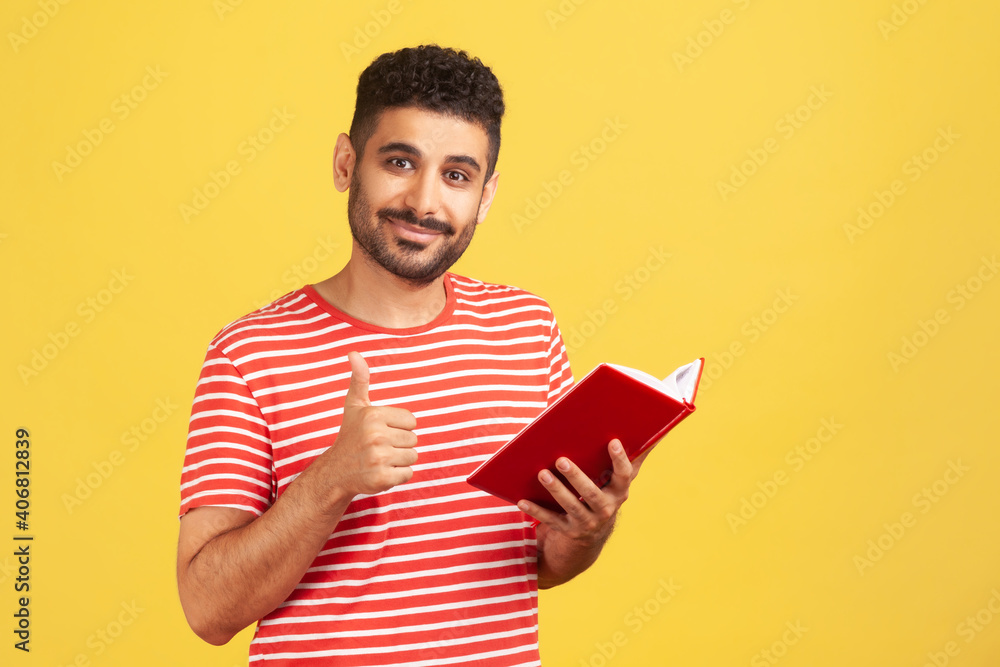 Happy satisfied bearded man in red striped t-shirt showing thumbs up gesture holding and reading book, likes genre and plot. Indoor studio shot isolated on yellow background Happy satisfied bearded man in red striped t-shirt showing thumbs up gesture holding and reading book, likes genre and plot. Indoor studio shot isolated on yellow background