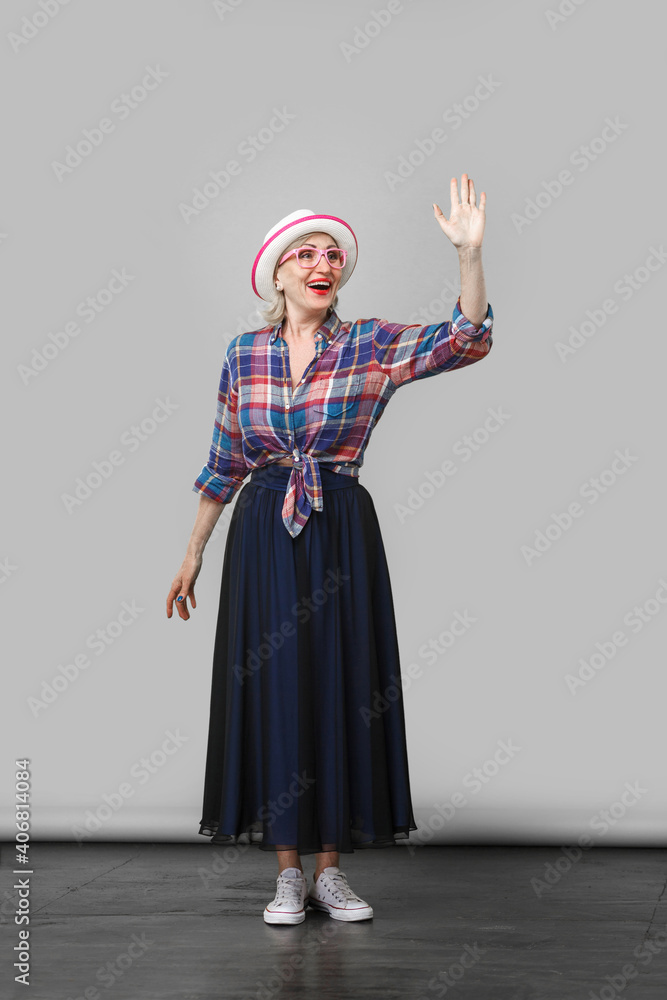 full length portrait of excited modern stylish mature woman in casual style with hat, eyeglasses standing, waving her hand and looking with toothy smile. indoor studio shot on gray wall background.