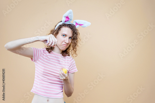Funny angry girl in bunny ears, striped pink t-shirt is boxing with eggs. Young curly woman is preparing for celebration. Happy easter spring concept. Carnival, seasonal party decor for holiday.