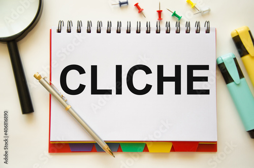 the word cliche is written on a white pad that lies on a white table near a magnifying glass of markers and multi-colored buttons photo