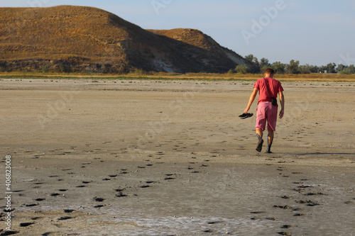 barefoot young man of the European race in a red t-shirt and pink shorts with slaps in hand goes away in the mud