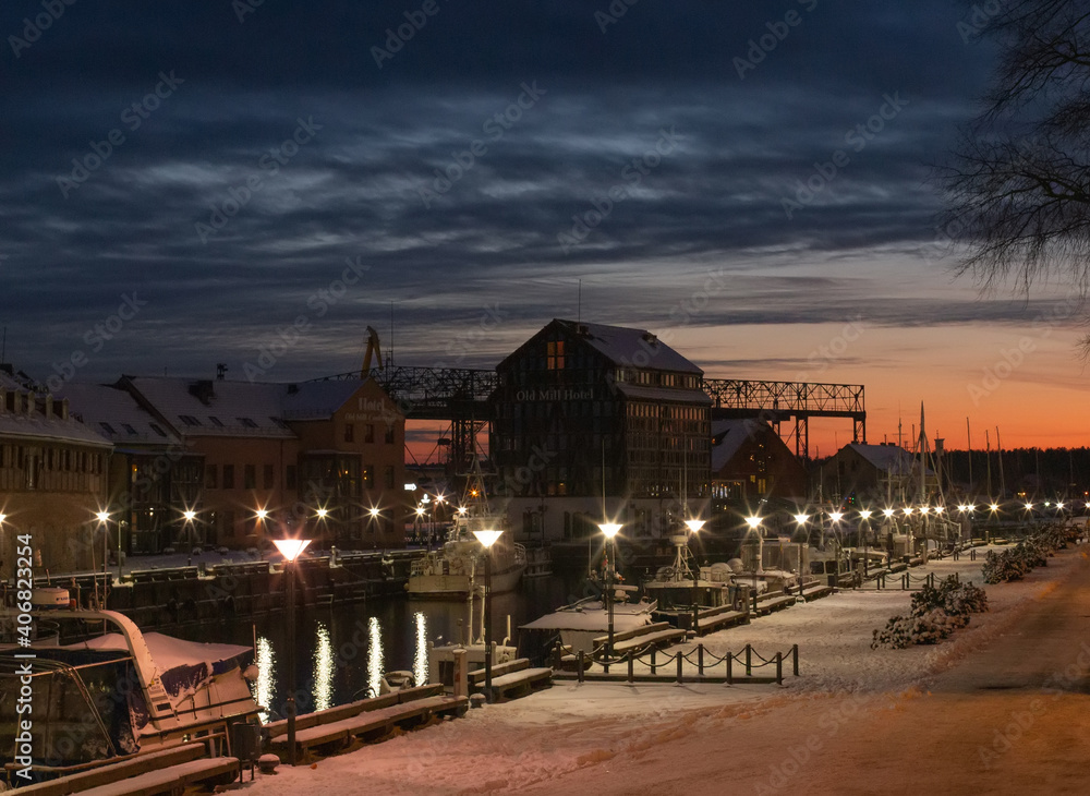 Klaipeda city, Lithuania in January. An old town, river and boats covered by winter snow, cold weather, empty streets. Beautiful red orange twilight sunset sky and street lights