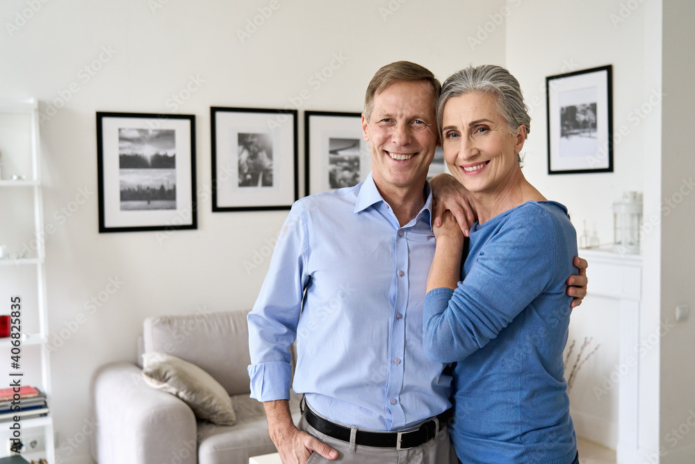 Fototapeta premium Happy senior mature 60s family couple hugging, looking at camera, standing in living room in modern apartment. Smiling satisfied middle aged husband and wife embracing posing for portrait at new home.