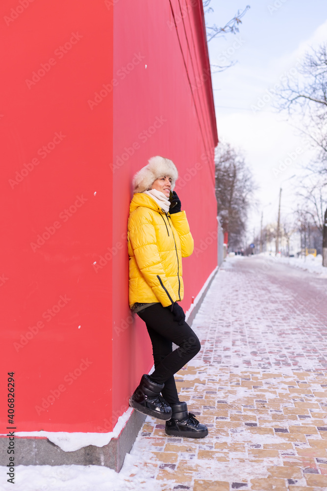 Happy young woman on a background of a red wall in warm clothes on a winter sunny day smiling and talking on the phone on a snowy city sidewalk Happy young woman on a background of a red wall in warm clothes on a winter sunny day smiling and talking on the phone on a snowy city sidewalk