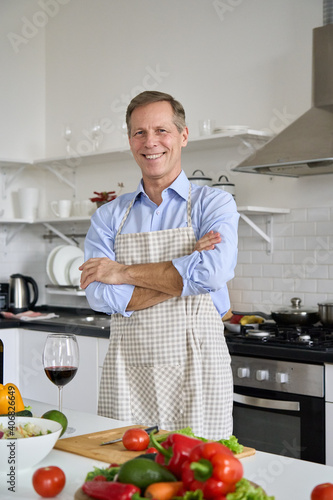 Happy middle aged single man wearing apron cooking at home. Smiling handsome 50s mature old chef preparing salad looking at camera standing in kitchen. Senior people healthy food concept, portrait.