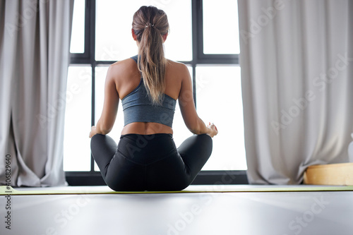 woman in sportive top and leggings practicing yoga at home sitting in lotus pose, practice meditation. rear view
