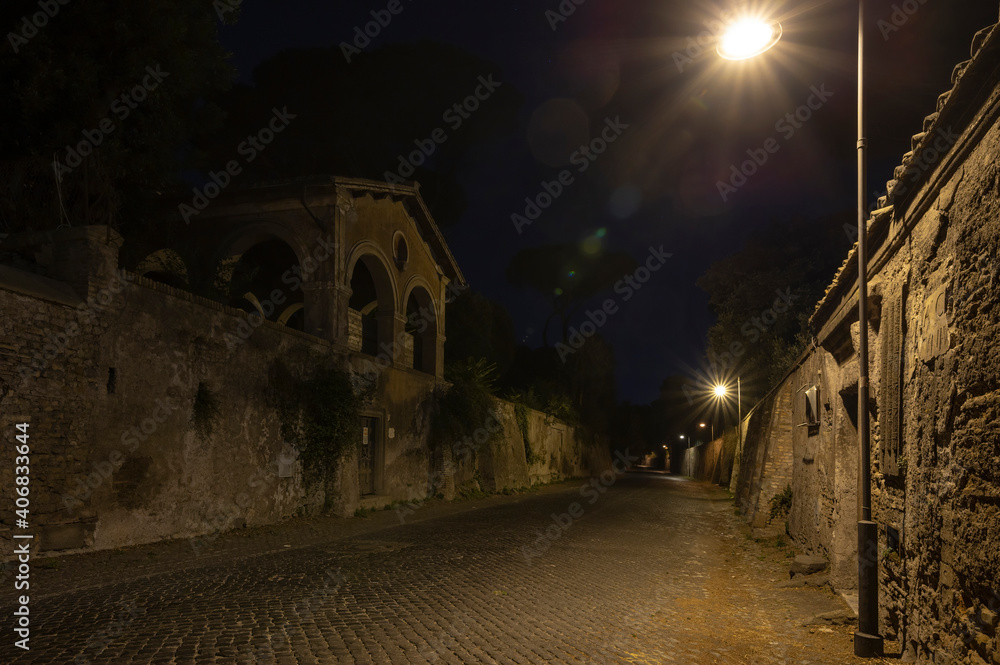 Via Appia Antica at night, beautiful detail of the lights of the street lamps, on the road near the Sepolcro degli Scipioni and the Porta di San Sebastiano, with the Aurelian Walls museum. Rome Italy