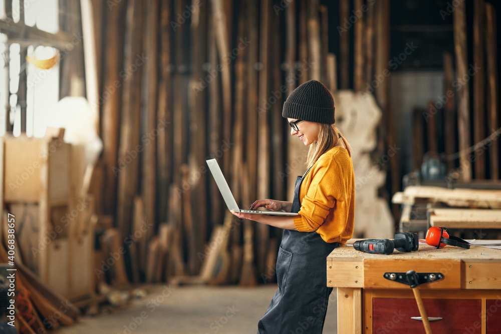 Fototapeta premium young female carpenter looks drawings on a laptop during a break in work in workshop