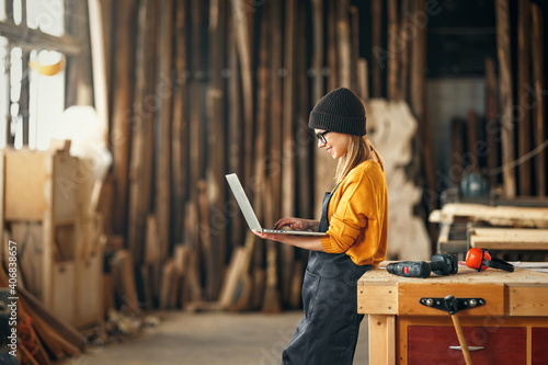 young female carpenter looks drawings on a laptop during a break in work in  workshop