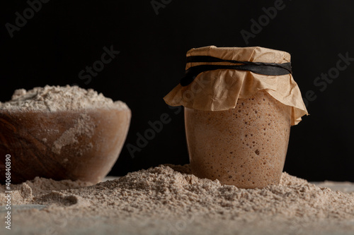 Homemade sourdough bread making ingredients featuring a bowl of flour and a sourdough starter culture in a glass cup covered with a brown paper on top. They all sit on flour covered kitchen countertop