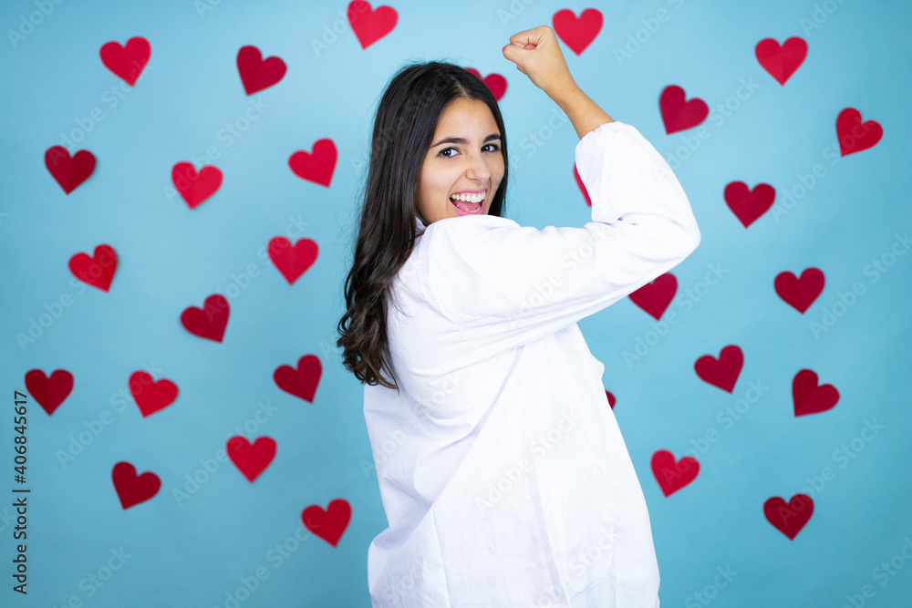 Young doctor woman wearing medical coat and stethoscope over blue background with red hearts feeling happy, satisfied and powerful, flexing fit and muscular biceps, looking strong after the gym Young doctor woman wearing medical coat and stethoscope over blue background with red hearts feeling happy, satisfied and powerful, flexing fit and muscular biceps, looking strong after the gym