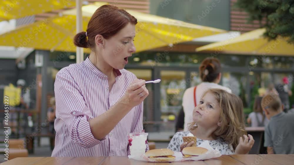 Young Mother And Little Girl In Cafe Eating Ice Cream. Mother Feeds Her Daughter With Spoon. Young Mother And Little Girl In Cafe Eating Ice Cream. Mother Feeds Her Daughter With Spoon.