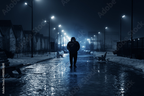 Silhouette of lonely person walks on dark foggy street illuminated with street lamps, blue toned.