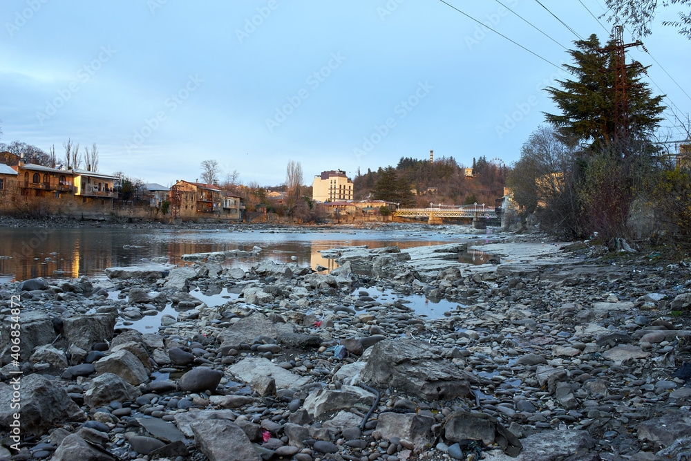 The banks of a river full of stones, with the view of the other bank full of old houses.  