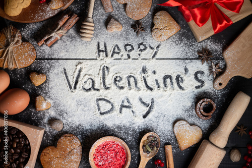 Happy Valentines Day written on flour. Gingerbread cookies, spices, coffee beans, baking supplies on black wood surface