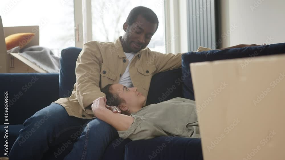 Young mixed ethnicity couple celebrating moving day in new home lying on sofa in lounge surrounded by removal boxes together - shot in slow motion Young mixed ethnicity couple celebrating moving day in new home lying on sofa in lounge surrounded by removal boxes together - shot in slow motion