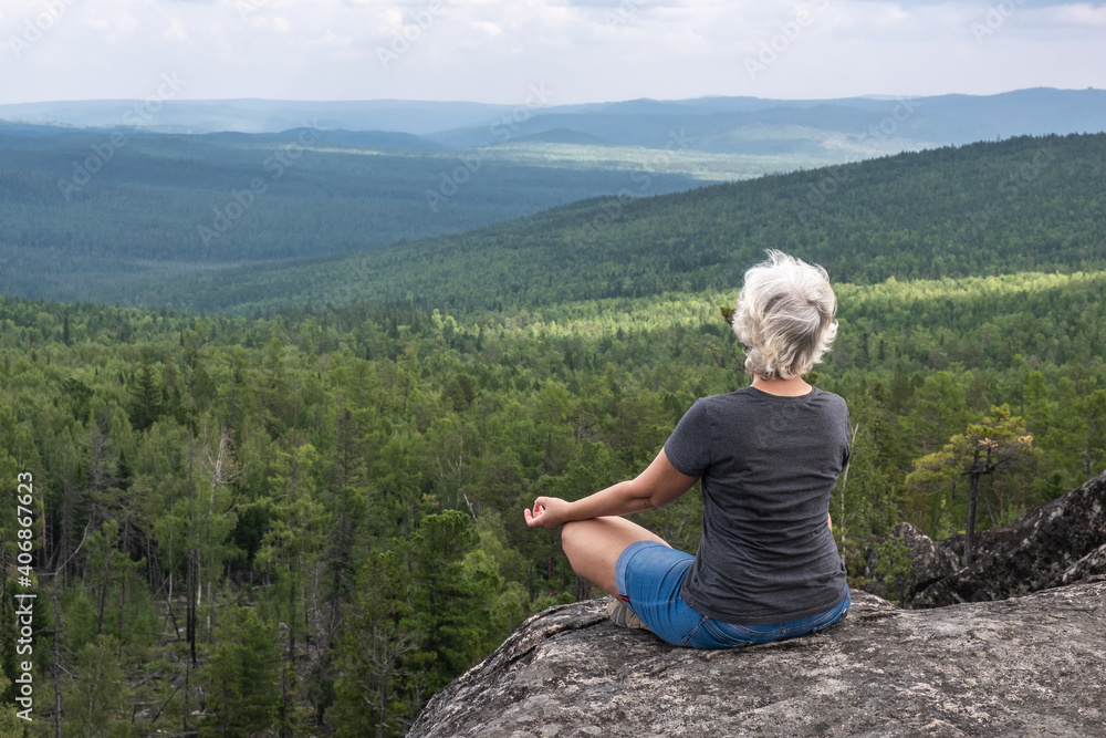 Naklejka premium A middle aged woman practices yoga and meditation on a mountain top with a stunning view of a forested, hilly valley.