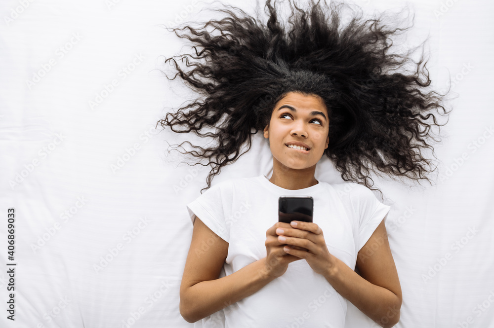 Wonderful curly woman is lying on the bed. Smiling African American girl use smartphone, think about shopping or searching new job, or planning vacation Wonderful curly woman is lying on the bed. Smiling African American girl use smartphone, think about shopping or searching new job, or planning vacation