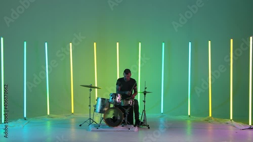 Expressive male musician plays drums in the studio against a background of multicolored neon tubes. A drummer with tattoos on his arms plays rock music with enthusiasm. Music concept. Slow motion. photo