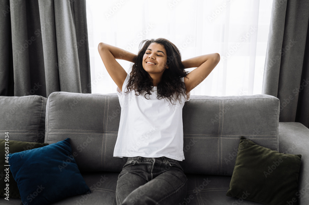 Satisfied beautiful African American girl sits on the sofa in living room. Happy young woman thinking about plans for weekends or dreaming about vacation and smiling Satisfied beautiful African American girl sits on the sofa in living room. Happy young woman thinking about plans for weekends or dreaming about vacation and smiling