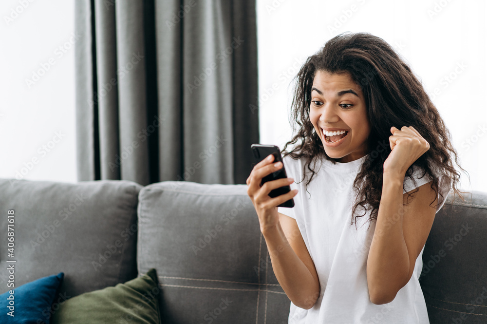 Fototapeta premium Overjoyed young woman use smartphone, receive a good message. Happy african american girl get a promotion on work or passing exams. Joyful female freelancer sit on the sofa, enjoy good results