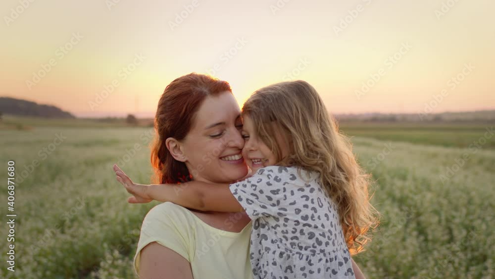 Young Mother Holding Her Daughter In Her Arms. They Are In The Middle Of The Field. They Are Both Smiling. Girl Kisses Mom. Young Mother Holding Her Daughter In Her Arms. They Are In The Middle Of The Field. They Are Both Smiling. Girl Kisses Mom.