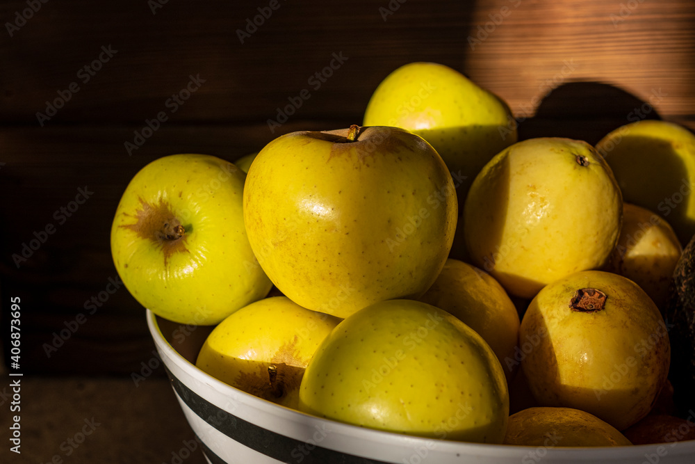 Manzanas amarillas y guayabas en un frutero blanco y negro, con luz de atardecer y madera de fondo